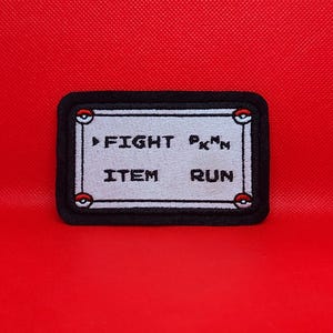 May include: A black and white embroidered patch with a white background and black text. The text reads "FIGHT, PKMN, ITEM, RUN". The patch has a black border and two red and white Pokeballs on either side.