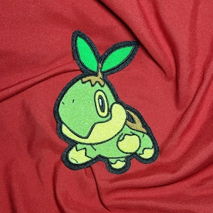 May include: Green and yellow embroidered patch of a cartoon turtle with a green leaf on its head. The turtle is standing on a red fabric background.