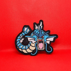May include: An embroidered patch featuring a cartoon dragon in blue and white with a black outline. The dragon has its mouth open wide and is facing the viewer. The patch is displayed against a red background.