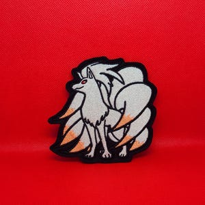 Peut inclure: Patch brodé blanc représentant un renard à neuf queues avec des pointes orange sur ses queues. Le renard a les yeux rouges et se tient sur un fond rouge.