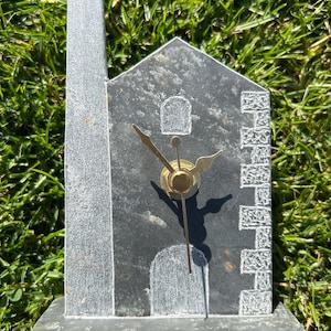 May include: A grey stone clock in the shape of a building with a chimney. The clock features a brass clock face and hands. The building has a door and window carved into the stone. The clock rests on a rectangular base.