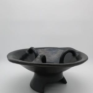 May include: A dark grey ceramic bowl with a wide, shallow basin and three curved handles. The bowl sits on a pedestal base with three feet. The design is minimalist and modern, suitable for decorative or functional use.