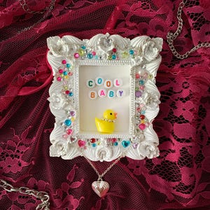 Puede incluir: Un marco de fotos blanco con gemas coloridas y las letras "COOL BABY". Dentro del marco hay un patito de goma amarillo. Un collar con colgante de corazón plateado cuelga debajo. El fondo es una tela de encaje rojo oscuro.