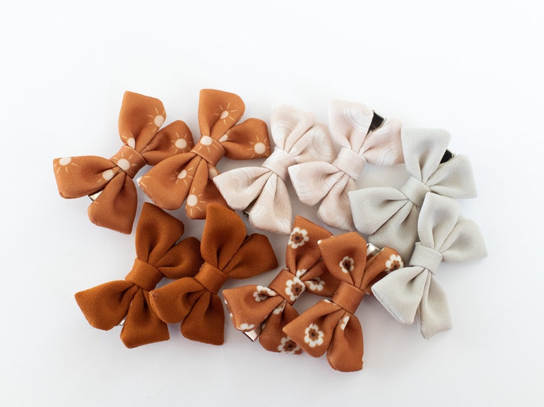 May include: A set of nine fabric hair bows in various patterns and colors. The bows are made of a soft, velvety fabric and feature a variety of designs, including sunbursts, floral prints, and solid colors. The bows are attached to alligator clips, making them easy to clip into hair.