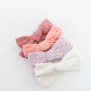 May include: Four fabric hair bows in different shades of pink, from dark pink to white. The bows are made of a delicate, eyelet fabric and are arranged in a stack.