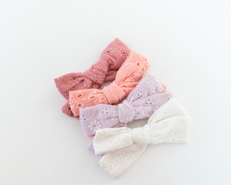 May include: Four fabric hair bows in different shades of pink, from dark pink to white. The bows are made of a delicate, eyelet fabric and are arranged in a stack.