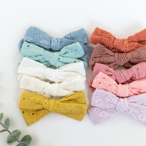 May include: A set of ten handmade hair bows in various pastel colors, featuring a delicate eyelet lace design. The bows are arranged in a stack, with the colors transitioning from light blue to lavender.
