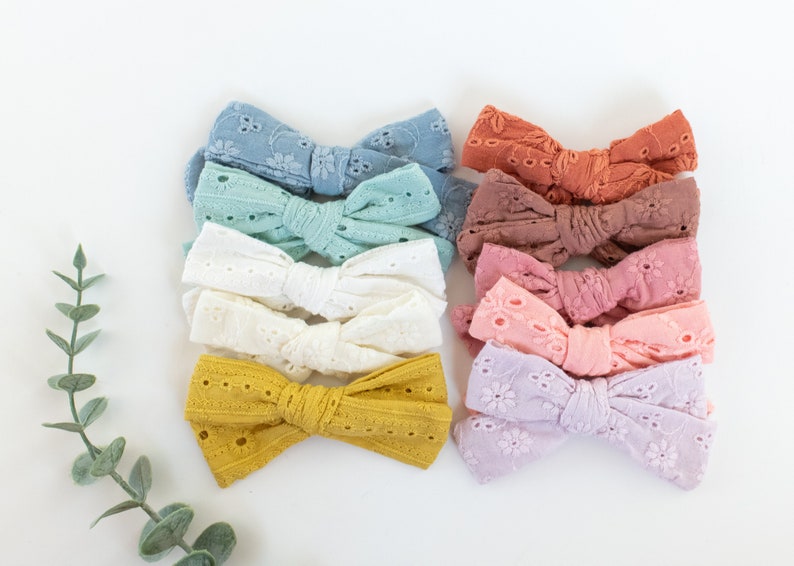 May include: A set of ten handmade hair bows in various pastel colors, featuring a delicate eyelet lace design. The bows are arranged in a stack, with the colors transitioning from light blue to lavender.