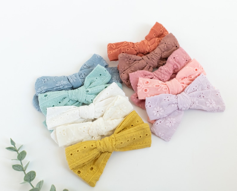 May include: A set of ten handmade fabric hair bows in various pastel colors, including light blue, green, white, yellow, pink, and purple. The bows are made of a delicate, eyelet fabric and are tied with a simple knot.