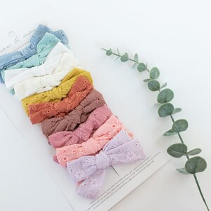 May include: A set of 10 colorful hair bows with eyelet lace detail. The bows are arranged in a stack, with the lightest color at the top and the darkest color at the bottom. The colors are light blue, teal, white, yellow, orange, red, pink, light pink, purple, and lavender.