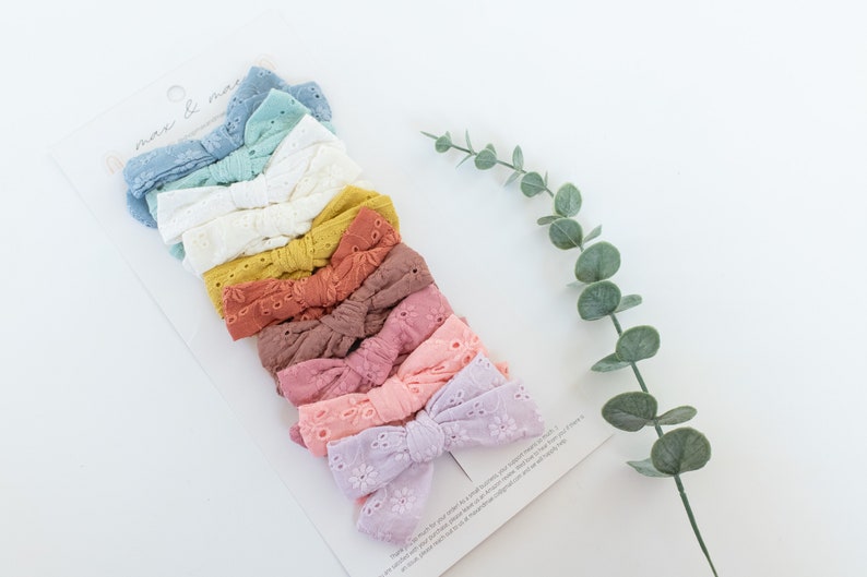 May include: A set of 10 colorful hair bows with eyelet lace detail. The bows are arranged in a stack, with the lightest color at the top and the darkest color at the bottom. The colors are light blue, teal, white, yellow, orange, red, pink, light pink, purple, and lavender.