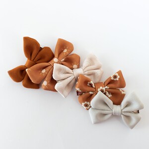 May include: Four fabric hair bows in a variety of patterns. The bows are in shades of brown and white. The bows are arranged in a row, with the largest bow at the top and the smallest bow at the bottom.