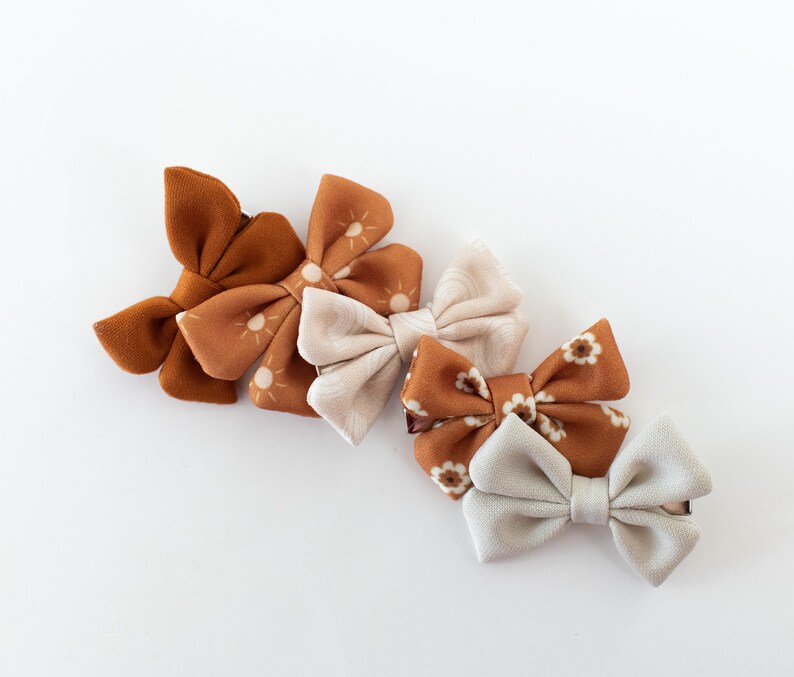 May include: Four fabric hair bows in a variety of patterns. The bows are in shades of brown and white. The bows are arranged in a row, with the largest bow at the top and the smallest bow at the bottom.