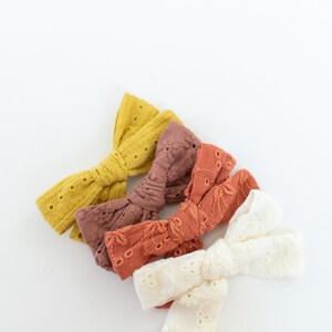May include: Three fabric hair bows in different colors. The bows are made of a soft, textured fabric with a delicate lace pattern. The colors are a light yellow, a light brown, and a white.