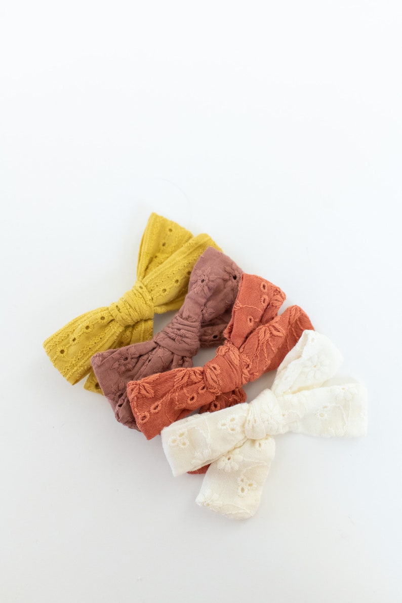May include: Three fabric hair bows in different colors. The bows are made of a soft, textured fabric with a delicate lace pattern. The colors are a light yellow, a light brown, and a white.