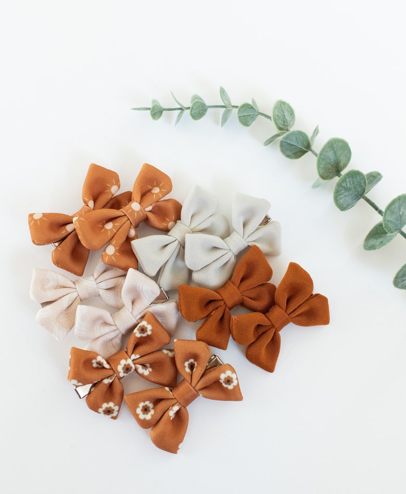 May include: A set of eight hair bows in two different patterns. Four bows are a light brown with a white sun pattern and four bows are a darker brown with a white flower pattern.