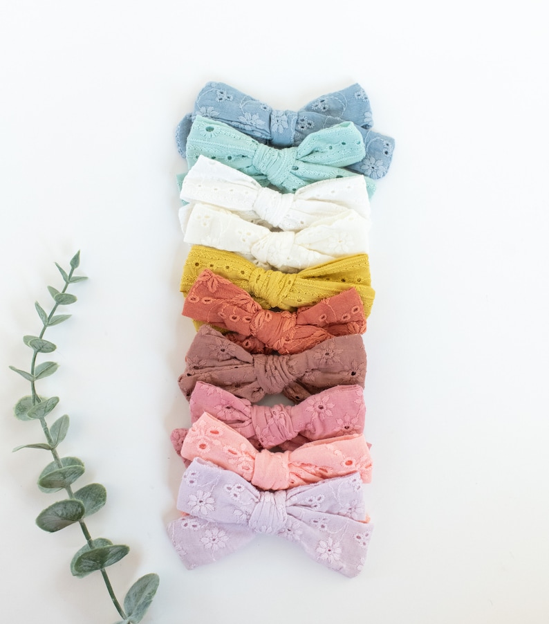 May include: A set of ten hair bows in various pastel colors, including light blue, white, yellow, orange, brown, pink, and purple. The bows are made of a delicate, eyelet fabric and are arranged in a vertical stack.