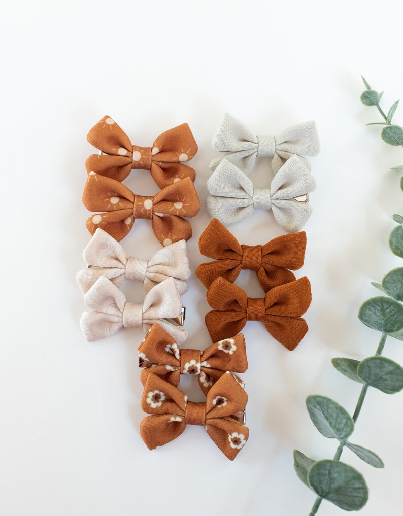 May include: Set of 8 brown and white fabric hair bows with a sun and floral print. The bows are attached to alligator clips.