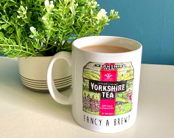 Può includere: Una tazza in ceramica bianca con un manico nero. La tazza presenta un'illustrazione colorata di una scatola di tè con il testo "Yorkshire Tea" e la frase "Fancy a Brew?" stampata sul fondo.