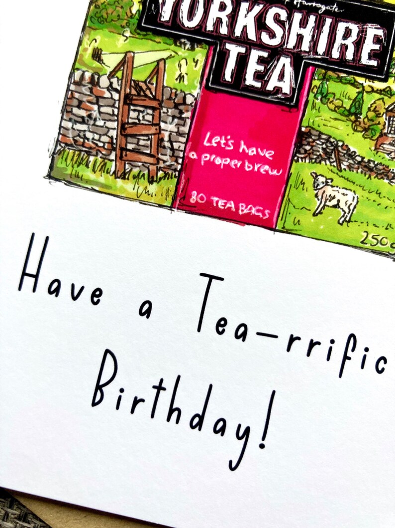 May include: A white card with black handwritten text that says "Have a Tea-rrific Birthday!"  The card has a small image of a tea box with a green and brown countryside scene and the words "Yorkshire Tea" in black and white. The tea box has a pink section with the words "Let's have a proper brew" and "80 Tea Bags" in white.
