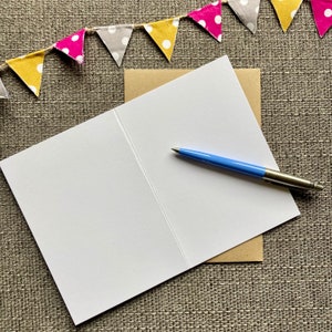 May include: A blank white card with a brown envelope and a blue pen. The card is folded in half and the envelope is partially covering the card. The pen is lying on top of the card.