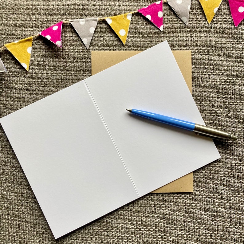 May include: A blank white card with a brown envelope and a blue pen. The card is folded in half and the envelope is partially covering the card. The pen is lying on top of the card.