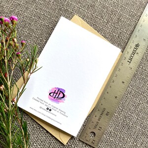 May include: A white card with a purple and pink watercolor design of the letters "HD" in a circle. The text "Printed from an original drawing" and "Created from start to finish in Yorkshire" is printed below the design. The text "@hddesignsyorkshire" and "www.etsy.com/shop/hddesignsyorkshire" is printed below the text. The card is sitting on a brown envelope.