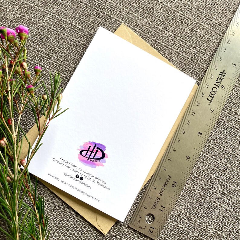 May include: A white card with a purple and pink watercolor design of the letters "HD" in a circle. The text "Printed from an original drawing" and "Created from start to finish in Yorkshire" is printed below the design. The text "@hddesignsyorkshire" and "www.etsy.com/shop/hddesignsyorkshire" is printed below the text. The card is sitting on a brown envelope.