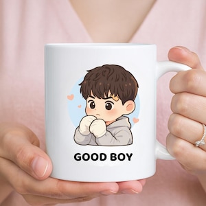 May include: White ceramic mug featuring a cartoon illustration of a boy with brown hair, wearing a gray hoodie and white gloves. The text "GOOD BOY" is printed in black below the illustration. Two small pink hearts are on the background.