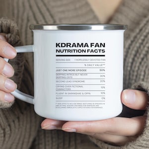 以下が含まれることがあります： ステンレス製の蓋とハンドルが付いた白いエナメル製のキャンプマグカップ。マグカップには、「Kdrama Fan Nutrition Facts」というテキストと、「Just One More Episode」や「Crying Over Fictional Characters」などの活動の「1日の摂取量」のパーセンテージのリストが書かれた白黒のグラフィックが描かれています。