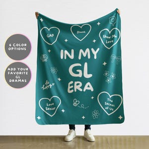 Pode incluir: Cobertor turquesa com texto e ilustrações brancas. O cobertor diz "IN MY GL ERA" com desenhos de coração, flor e estrela. O texto adicional inclui "GAP", "Blank", "Show Me Love", "Love Senior" e "The Secret of Us".