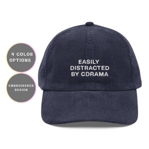 Puede incluir: Una gorra de béisbol de pana azul marino con texto bordado en blanco que dice "EASILY DISTRACTED BY DRAMA".