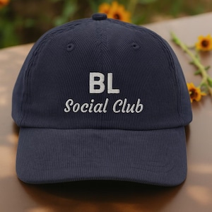 Könnte beinhalten: Marineblaue Cord-Baseballkappe mit dem weißen gestickten Text "BL Social Club". Die Kappe hat einen gebogenen Schirm und einen Knopf oben. Gelbe Blumen sind im Hintergrund zu sehen.
