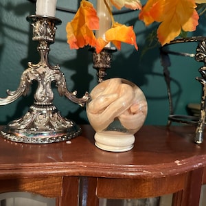 May include: A clear glass orb containing a coiled, pale-coloured object, possibly a preserved specimen. The orb sits on a white base. The background includes ornate silver candle holders, white candles, and orange and yellow artificial leaves.