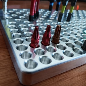 May include: A silver metal tool organizer with multiple holes, holding various screwdriver bits. The bits are in different colors, including red and black. The organizer is on a wooden surface.