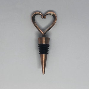 May include: A copper-coloured metal bottle stopper featuring a heart-shaped handle at the top. The stopper has a tapered, pointed base and a black ribbed silicone seal designed to fit securely into the neck of a bottle for sealing purposes.