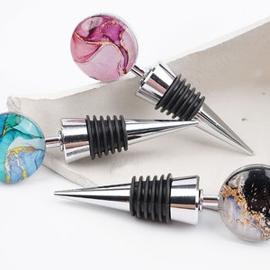 May include: Three wine bottle stoppers with different coloured tops. The tops are round and feature abstract marble patterns in pink, blue, and black with gold accents.