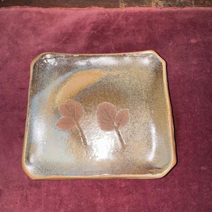 May include: A square ceramic plate with a brown and gold glaze. Two brown leaf designs are embossed on the plate's surface. The plate has a slightly irregular shape and a warm, earthy aesthetic.