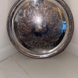 May include: A round silver-colored serving tray featuring a polished center and an intricate embossed floral pattern. The tray has a raised, textured rope-style border along the outer edge, providing a decorative finish to the metallic surface.