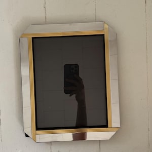 May include: A rectangular wall mirror featuring a reflective glass surface framed by alternating panels of smooth silver-toned mirror and vertical gold-toned ribbed metal accents. The design includes beveled edges and a dark central reflection.