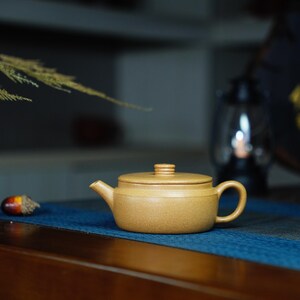 May include: A small, light brown teapot with a lid and a handle. The teapot is sitting on a blue mat.