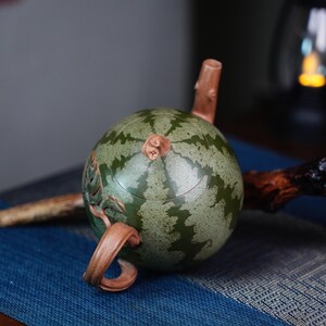 May include: A green and brown ceramic teapot shaped like a watermelon. The teapot has a carved handle and spout.