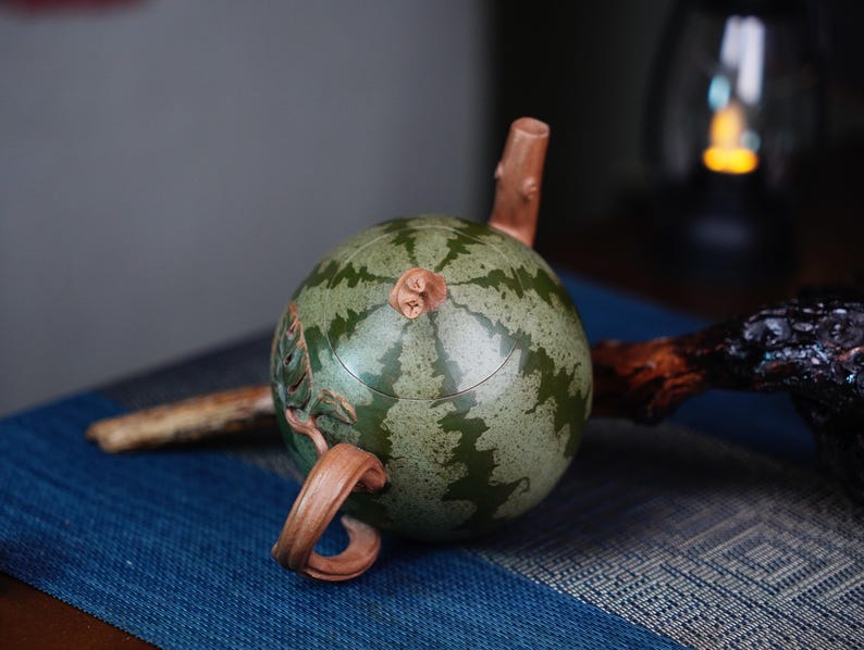 May include: A green and brown ceramic teapot shaped like a watermelon. The teapot has a carved handle and spout.