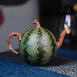 May include: A green and brown ceramic teapot shaped like a watermelon. The teapot has a handle and a spout.