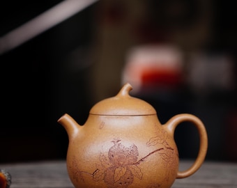 May include: A small, tan ceramic teapot with a rounded body, a curved handle, and a spout. The teapot features a detailed brown illustration of a pomegranate and leaves. The lid has a small knob. The teapot sits on a wooden surface.