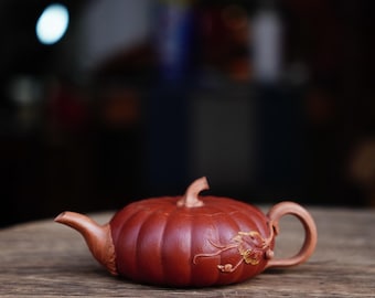 May include: A red clay teapot shaped like a pumpkin with a carved floral design. The teapot is sitting on a wooden surface.