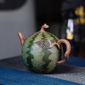 May include: A green and brown ceramic teapot shaped like a watermelon with a leafy design. The teapot has a spout and a handle.