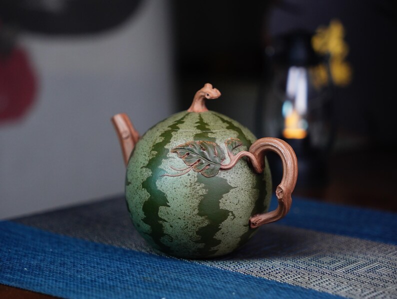 May include: A green and brown ceramic teapot shaped like a watermelon with a leafy design. The teapot has a spout and a handle.