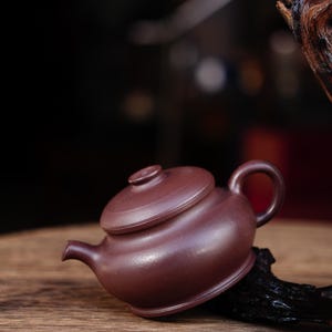 以下が含まれることがあります： 丸みを帯びたボディと蓋が付いた、濃い赤色の粘土製のティーポット。ティーポットは木製の表面の上に置かれています。