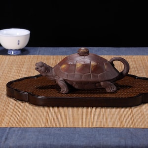 May include: A brown ceramic teapot shaped like a turtle, with a lid and a spout. The teapot is sitting on a wooden base with a brown and black design. The teapot is on a brown woven mat.